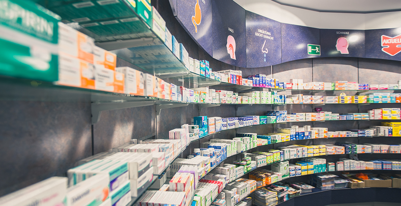 Innenaufnahme einer modernen Apotheke mit Regalen voller Gesundheitsprodukte und Medikamenten. Die Regale sind ordentlich organisiert und bieten eine breite Auswahl an Produkten, von Schmerzmitteln bis hin zu Hautpflegeprodukten. Der Raum ist gut beleuchtet, was für eine einladende Atmosphäre sorgt.