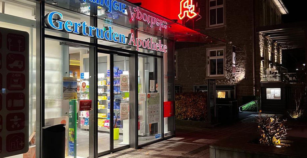 Außenansicht der Gertruden Apotheke bei Nacht. Die Glasfassade ist hell beleuchtet, über dem Eingang leuchtet das rote Apotheken-A. Das Schild „Gertruden Apotheke“ ist in Blau und Rot beleuchtet. Rechts ist ein historisches Gebäude aus Stein zu sehen.