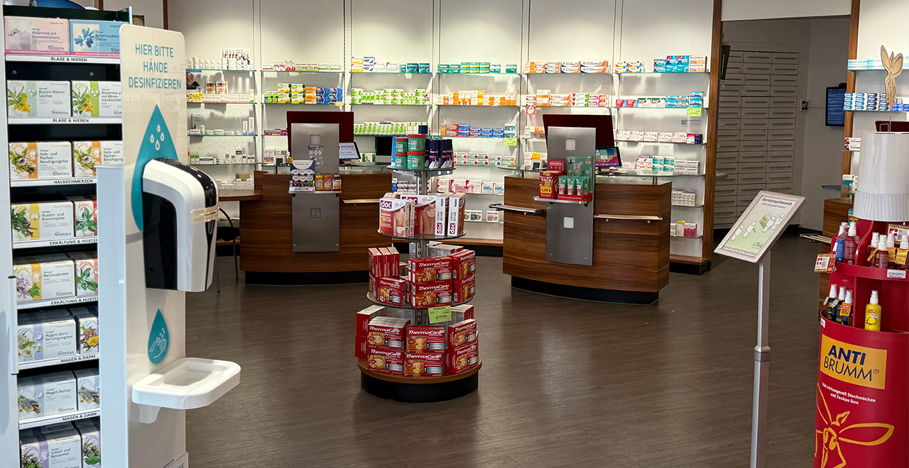 Innenaufnahme der Apotheke mit zentral platziertem runden Verkaufsständer mit Gelenkgelen und Wärmepflastern. Im Hintergrund zwei Holzverkaufstresen mit Monitoren und zahlreiche Produktregale mit Medikamenten und Pflegeprodukten. Rechts ein roter Werbeaufsteller für „ANTI BRUMM“.