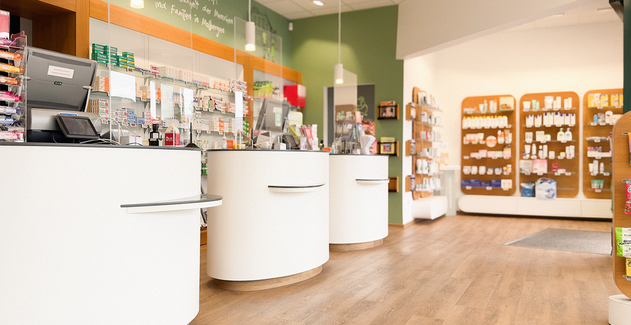 Heller, modern eingerichteter Verkaufsbereich einer Apotheke mit weißen, runden Beratungstheken, Medikamentenregalen im Hintergrund und Holzfußboden.
