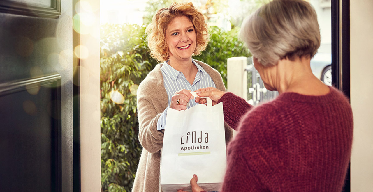 Eine ältere Frau bekommt an der Haustür eine Apotheken-Tüte von einer lächelnden Botin überreicht.