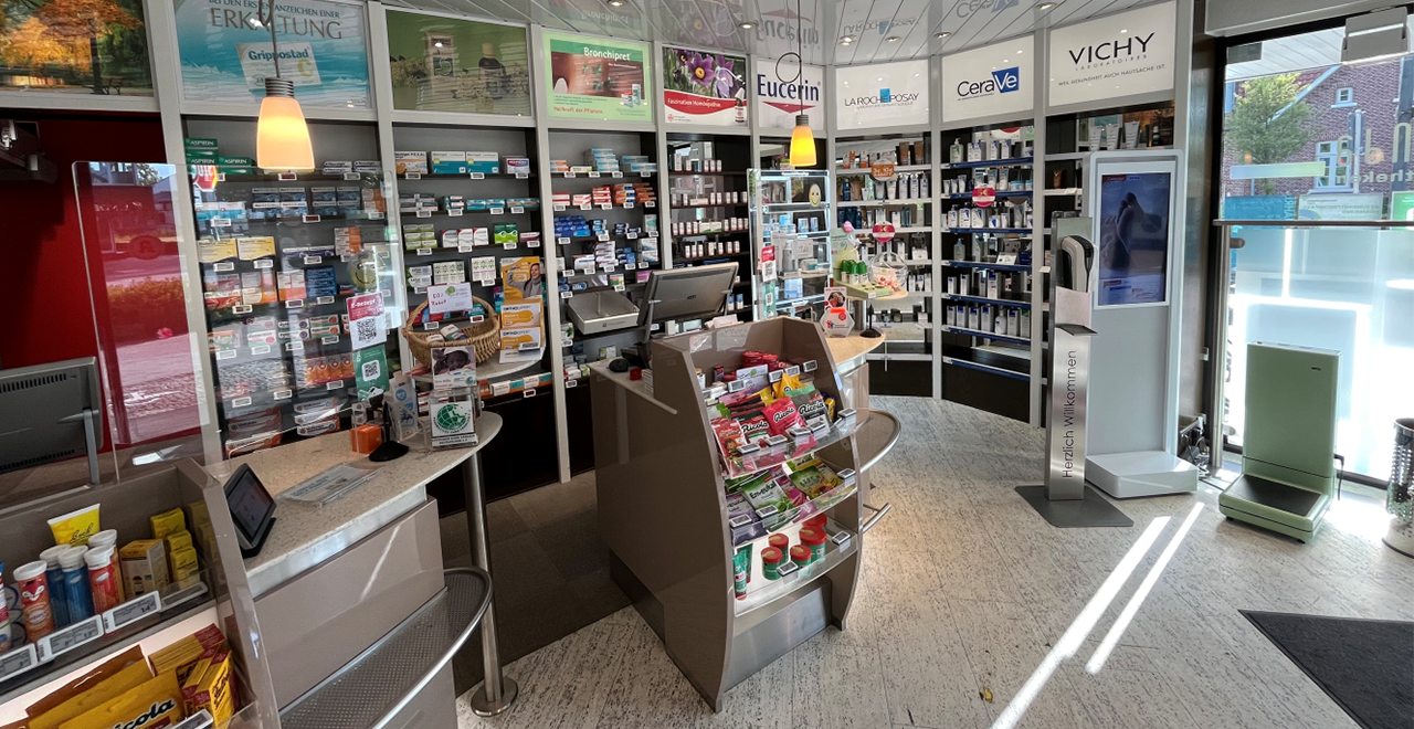 Apotheken-Innenraum mit Medikamentenregalen, Kassenplätzen und einem klar strukturierten Verkaufsraum. Ein Durchgang im Hintergrund führt zu einem Nebenraum oder Büro.