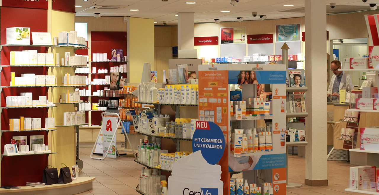 Verkaufsraum einer Apotheke mit Regalen und Präsentationsständern für Medikamente und Pflegeprodukte; im Hintergrund eine Person im weißen Kittel.