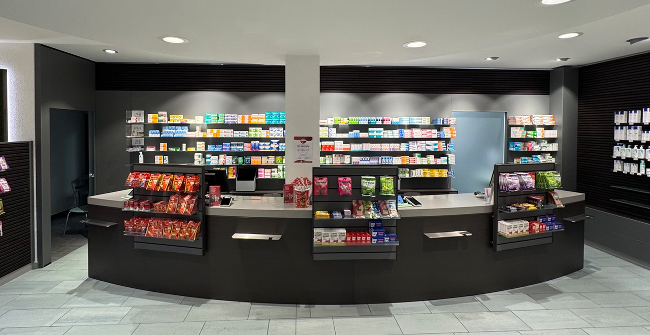 Innenraum einer modernen Apotheke mit Verkaufstresen, Regalen im Hintergrund und frei zugänglichen Produkten im Vorderbereich.