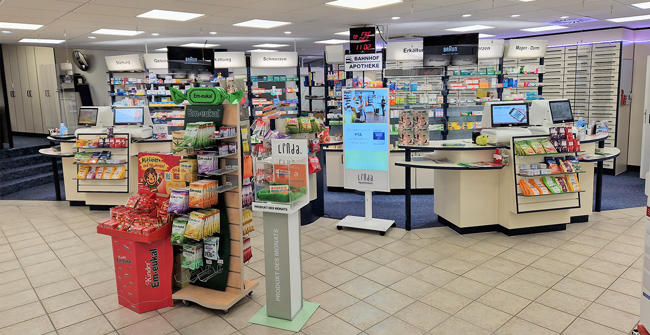 Innenansicht einer Apotheke mit moderner Einrichtung. Im Vordergrund stehen Verkaufsdisplays mit Em-eukal Bonbons und weiteren Gesundheitsprodukten. Dahinter befinden sich zwei Kassentresen mit Monitoren, sowie Regale mit Medikamenten. Über den Regalen sind Schilder mit Kategorien wie „Erkältung“, „Schmerzen“ und „Magen-Darm“ zu sehen. In der Mitte steht ein digitaler Werbebildschirm.