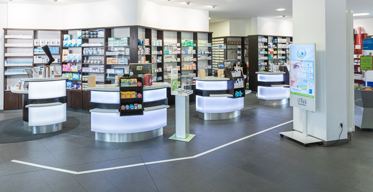 Innenansicht einer modernen Apotheke mit mehreren beleuchteten Beratungstheken, hinter denen gut sortierte Regale mit Medikamenten und Produkten zu sehen sind. Ein digitales Informationsdisplay steht im Vordergrund.