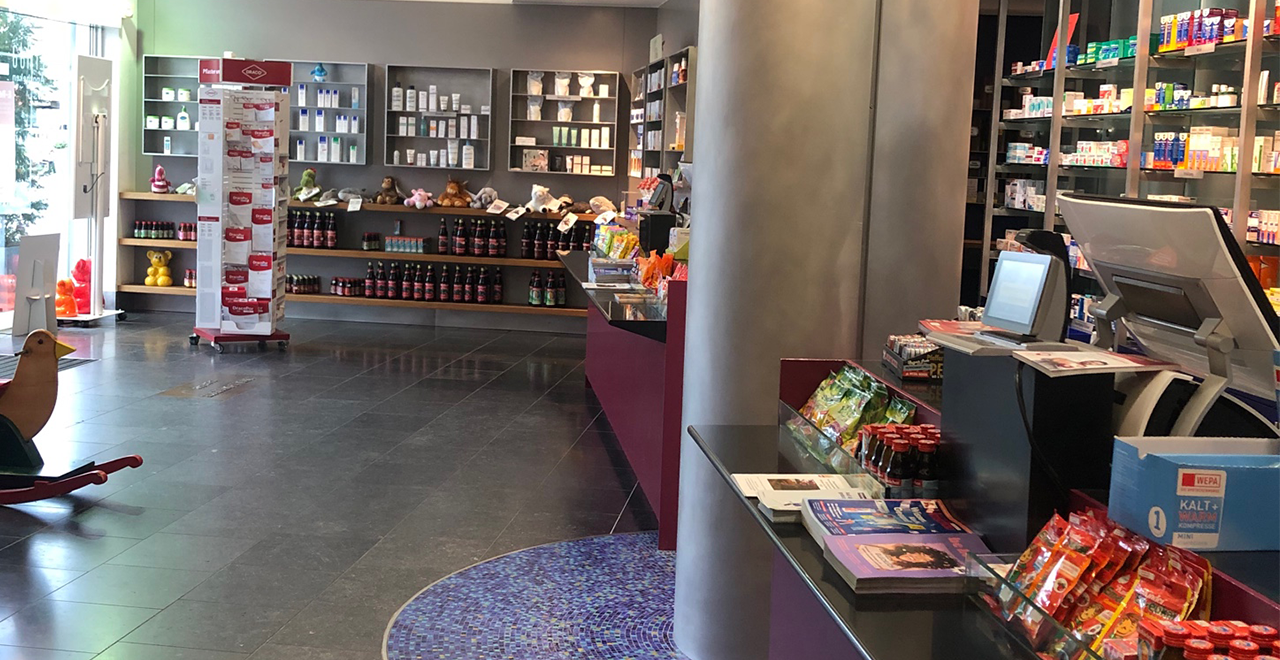 Heller Verkaufsraum einer Apotheke mit Regalen voller Medikamente und Pflegeprodukte, Theke mit Kasse, Werbematerial und kleinen Artikeln, ebenerdiger Zugang ohne Stufen.