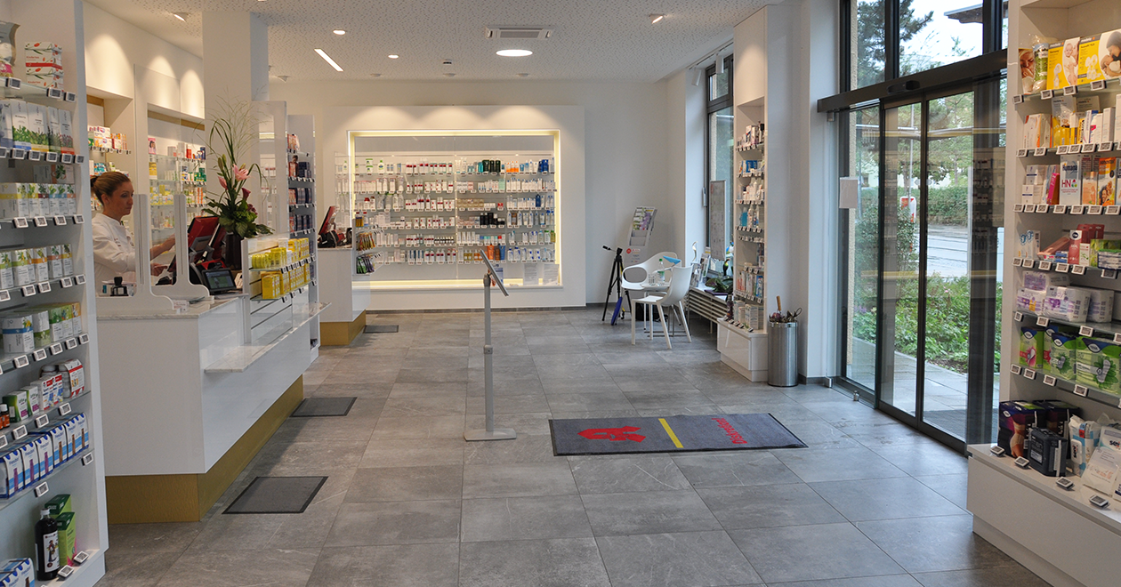 Helle, moderne Innenansicht einer Apotheke mit großzügigem Eingangsbereich. Links eine Mitarbeiterin in weißem Kittel hinter einer Beratungstheke mit Schutzscheibe. Die Verkaufsfläche ist aufgeräumt, mit eleganten Regalen an den Wänden, gefüllt mit Arzneimitteln und Pflegeprodukten. Rechts eine automatische Glastür mit Blick ins Grüne. Im Hintergrund ein kleiner Wartebereich mit zwei weißen Stühlen und einem Tisch.