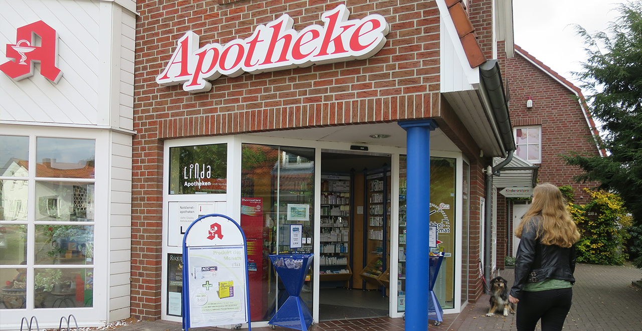 Eingang einer Apotheke mit rotem Schriftzug, einer Frau mit Hund davor und einem Werbeaufsteller neben der Tür.