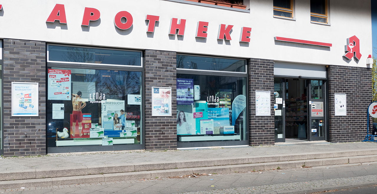 Außenansicht einer deutschen Apotheke mit dem großen roten Schriftzug „APOTHEKE“ über dem Eingang. Die Fenster sind mit zahlreichen Postern und Werbematerialien dekoriert, im Schaufenster stehen Produktpräsentationen. Der Eingangsbereich besteht aus zwei automatischen Glastüren. Die Fassade ist modern mit dunklen Klinkersteinen gestaltet.