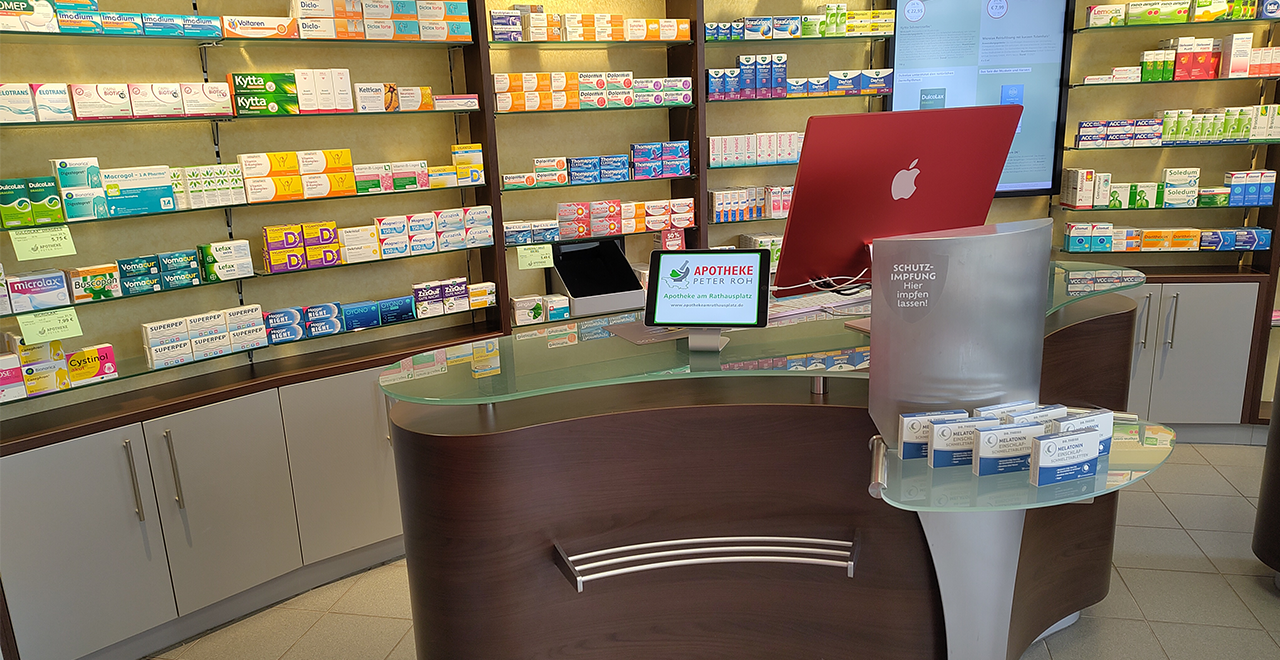 Innenansicht der Apotheke. Ein weiterer Blick auf die Regale mit Arzneimitteln und Produkten, die ordentlich auf den Wänden präsentiert sind. Im Vordergrund befindet sich ein moderner Empfangstresen mit einem Computer. Es gibt auch digitale Bildschirme, die Informationen anzeigen.
