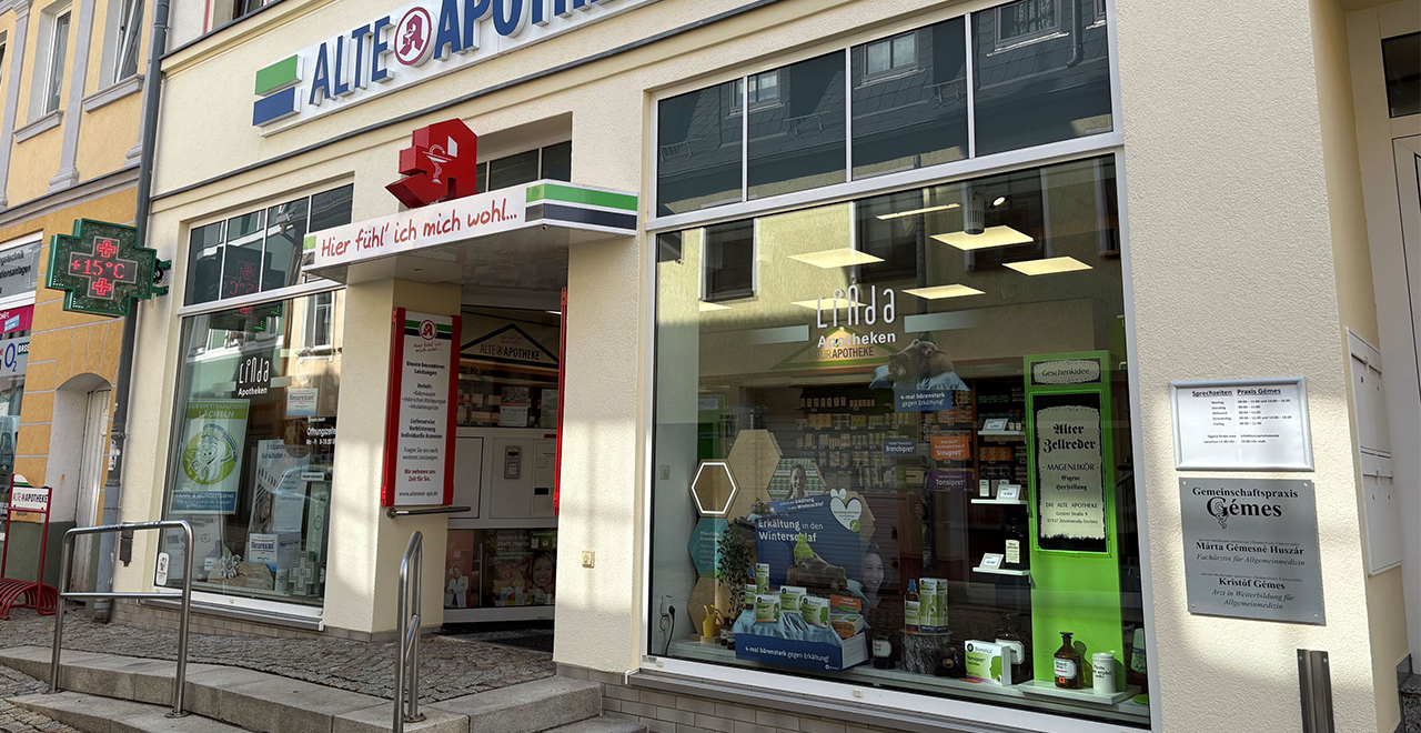 Frontale Gesamtansicht der Alten Apotheke mit zwei großen Schaufenstern und einem mittigen Eingang. Über dem Eingang befindet sich ein rotes Apothekenzeichen und ein Vordach mit der Aufschrift „Hier fühl’ ich mich wohl...“. In den Schaufenstern befinden sich Werbeposter und Produktpräsentationen. Rechts neben der Tür ist ein Praxisschild einer Arztpraxis angebracht.