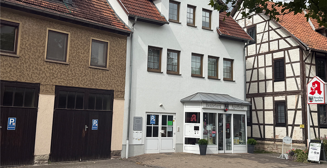 Die Henneberg Apotheke von der Straße aus, mit der weißen Fassade und einem großen Schild mit einem roten Hahn-Logo. Das Geschäft befindet sich zwischen zwei benachbarten Gebäuden, es sind Parkplätze mit blauen Schildern markiert.