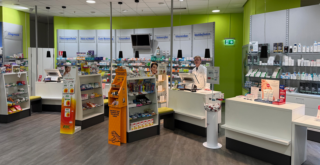 Heller Verkaufsraum einer Apotheke mit vielen Regalen voller Medikamente und Pflegeartikel. Im rechten Bereich steht ein Apotheker im weißen Kittel hinter einer Kasse. Mehrere freistehende Ständer mit Produkten wie Pflastern, Vitaminen und Wellnessartikeln befinden sich im Vordergrund. Die Wände sind grün, die Beleuchtung modern und gleichmäßig.
