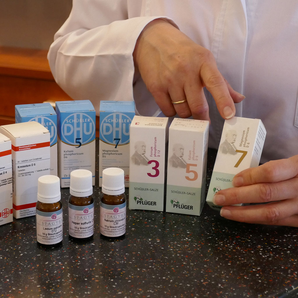 Aufreihung mehrerer unterschiedlicher Homöopathie- Arznei. Eine Frau in weißem Kitteln deutet auf eine Schachtel.