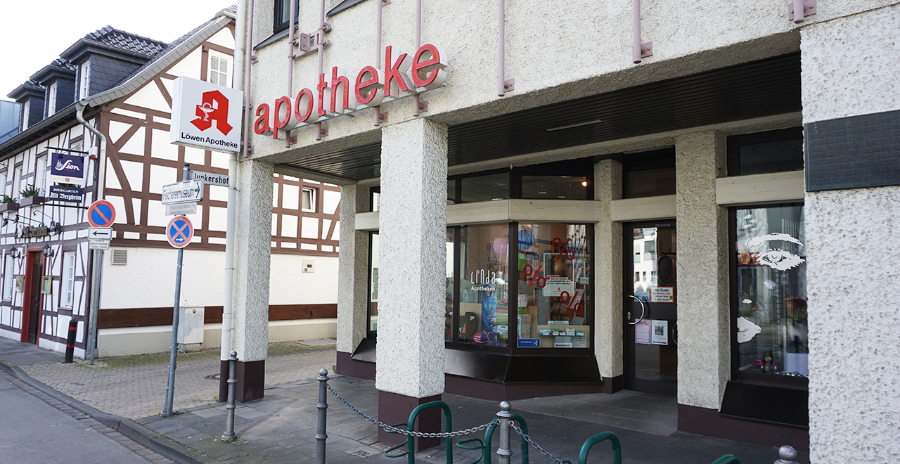 Außenansicht der Löwen Apotheke mit geparktem Apothekenfahrzeug davor. Auf dem Auto sind das Apothekenlogo und ein gelber Löwe abgebildet. Rechts neben der Apotheke befindet sich eine Filiale der Kreissparkasse. Ein Wegweiser zeigt Richtung Fischereimuseum.