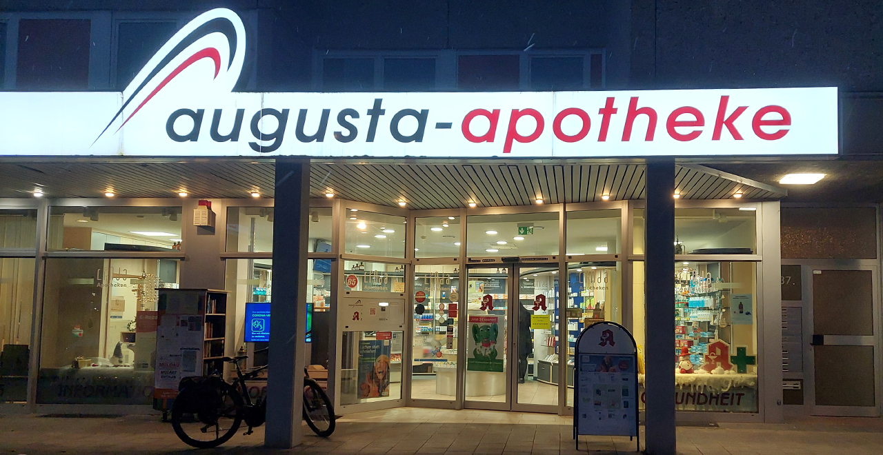 Außenansicht einer Augusta-Apotheke bei Nacht. Über dem Eingang leuchtet das Schild der Apotheke in weißer und roter Schrift. Der Eingang ist gut beleuchtet, und durch die Glaswände sind Regale mit verschiedenen Produkten sichtbar.