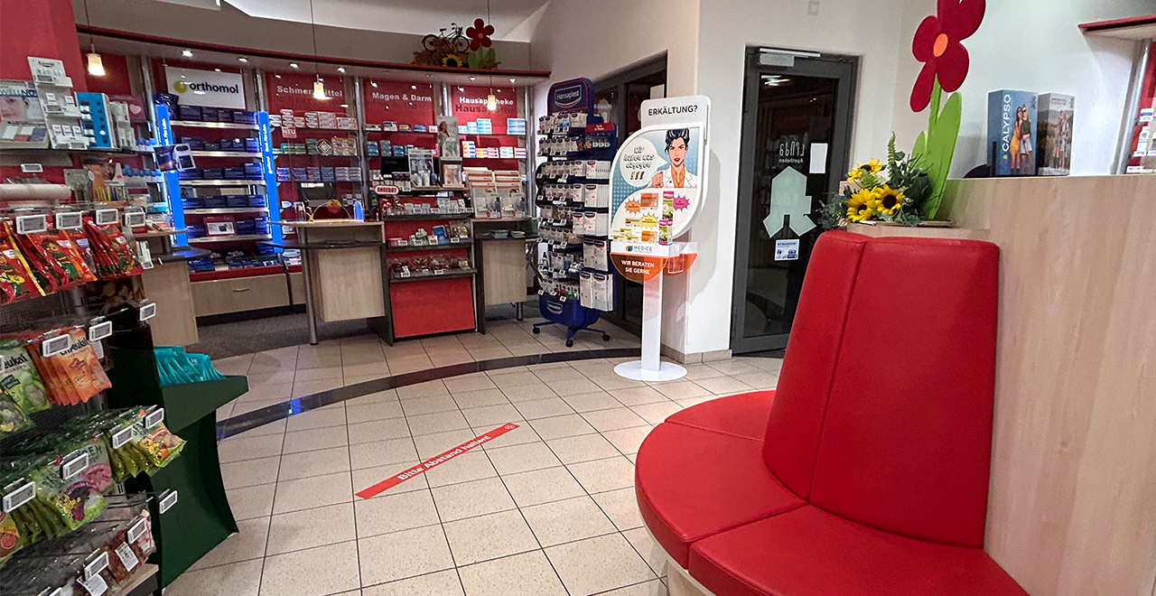 Innenraum einer Apotheke, im Vordergrund steht eine runde, rot gepolsterte Sitzbank. Links befinden sich Regale mit verschiedenen Produkten, darunter Nahrungsergänzungsmittel und Tees. Im Hintergrund ist der Verkaufsbereich mit roten Regalen und einer Theke zu sehen. Auf den Regalen stehen Medikamente und Gesundheitsprodukte. In der Mitte des Raumes ist ein Aufsteller mit Informationen zu Erkältungsprodukten.
