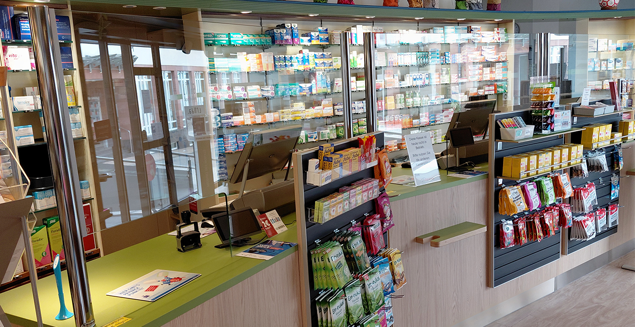Innenansicht der Adler-Apotheke mit Fokus auf die Verkaufsfläche und die hinteren Medikamentenregale. Die Sicht ist durch eine Glasscheibe gefiltert. Am vorderen Bereich stehen kleine Auslagen mit Bonbons und Ratgebern.