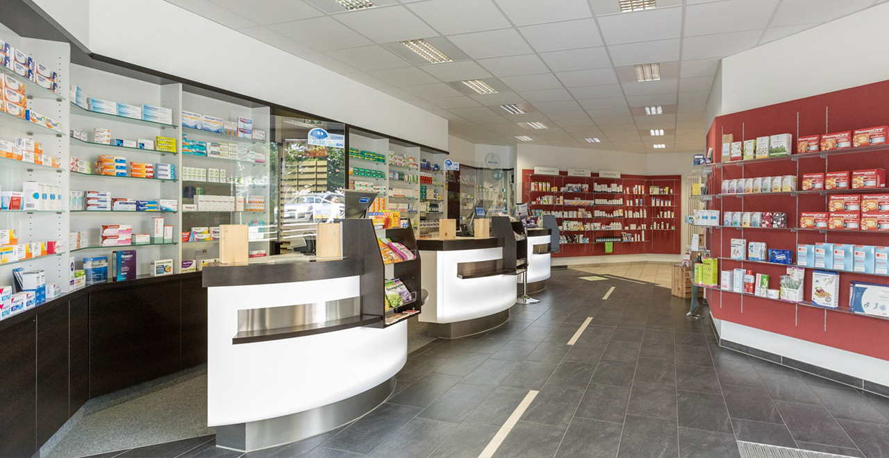 Eingangsbereich einer modernen Apotheke mit Glas-Schiebetüren, roter Backsteinfassade und einem Willkommensschild der "Finken-Apotheke". Im Inneren sind Regale mit Gesundheitsprodukten sichtbar.