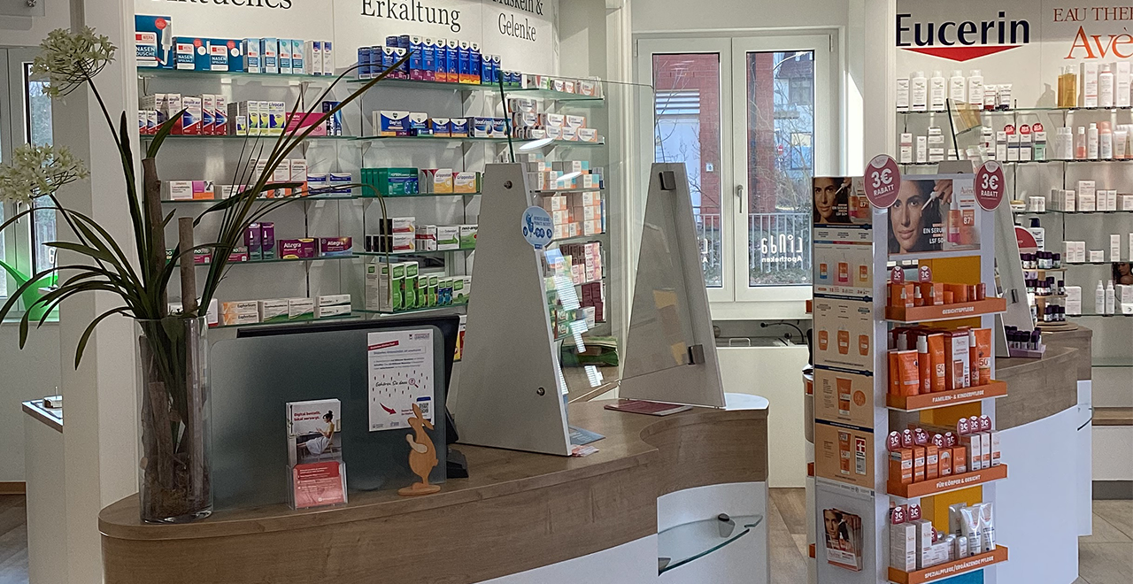 Innenansicht der Apotheke mit Verkaufstresen aus Holz und Plexiglas-Schutzwänden. Dahinter befinden sich Regale mit Medikamenten und Beschilderungen wie „Erkältung“. Rechts steht ein Verkaufsdisplay mit Hautpflegeprodukten der Marke Eucerin. Der Raum wirkt hell, sauber und gut organisiert.