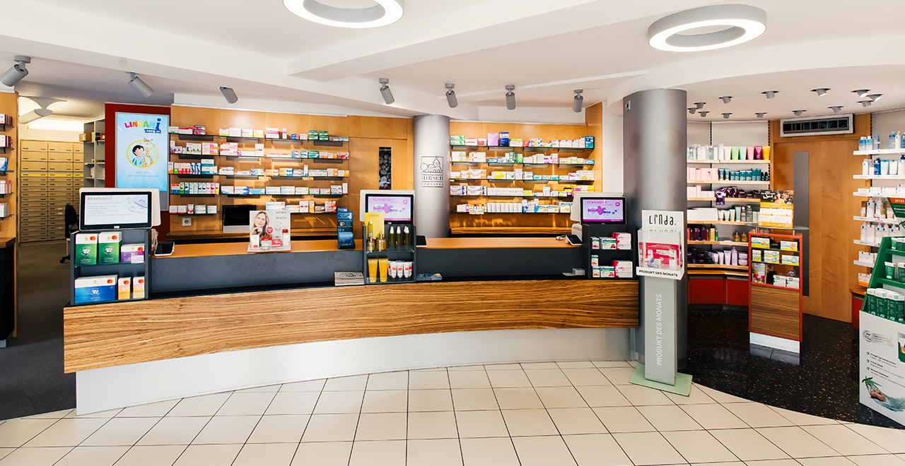 Innenansicht einer Apotheke mit einem langen Empfangstresen und Regalen, die mit Medikamenten und Gesundheitsprodukten bestückt sind. Zwei Tablets und Produktpräsentationen sind sichtbar.