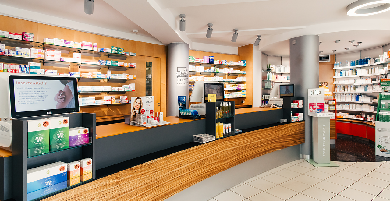 Weitere Innenansicht einer Apotheke mit einem Empfangstresen, der mit Produkten und Tablets ausgestattet ist. An den Wänden sind Regale mit Medikamenten und Gesundheitsprodukten zu sehen.