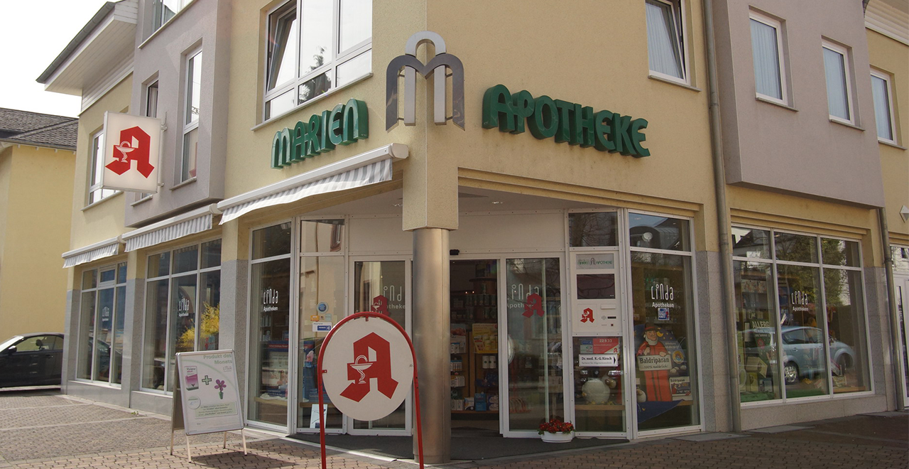 Der Eingang befindet sich an der Ecke des Gebäudes, mit dem Schriftzug „MARIEN APOTHEKE“ in grüner Schrift. Davor steht ein rundes Apotheken-A-Schild.