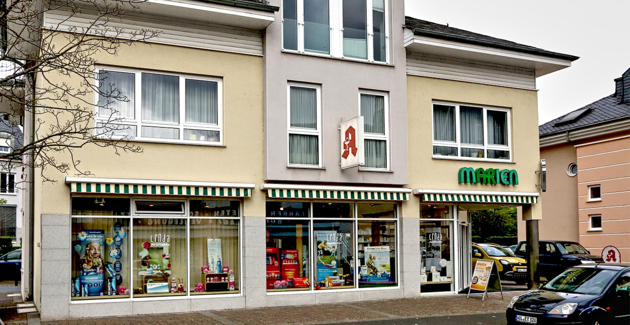 Eine moderne Apotheke in einem hellgelben Gebäude mit großen Schaufenstern und grün-weiß gestreifter Markise. Über dem Eingang steht „MARIEN“ in grüner Schrift, das rote Apotheken-A hängt an der Fassade.