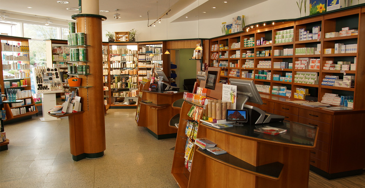 Heller, freundlicher Verkaufsraum einer Apotheke mit Holzregalen, Medikamenten, Kosmetikprodukten und zwei Kassen.
