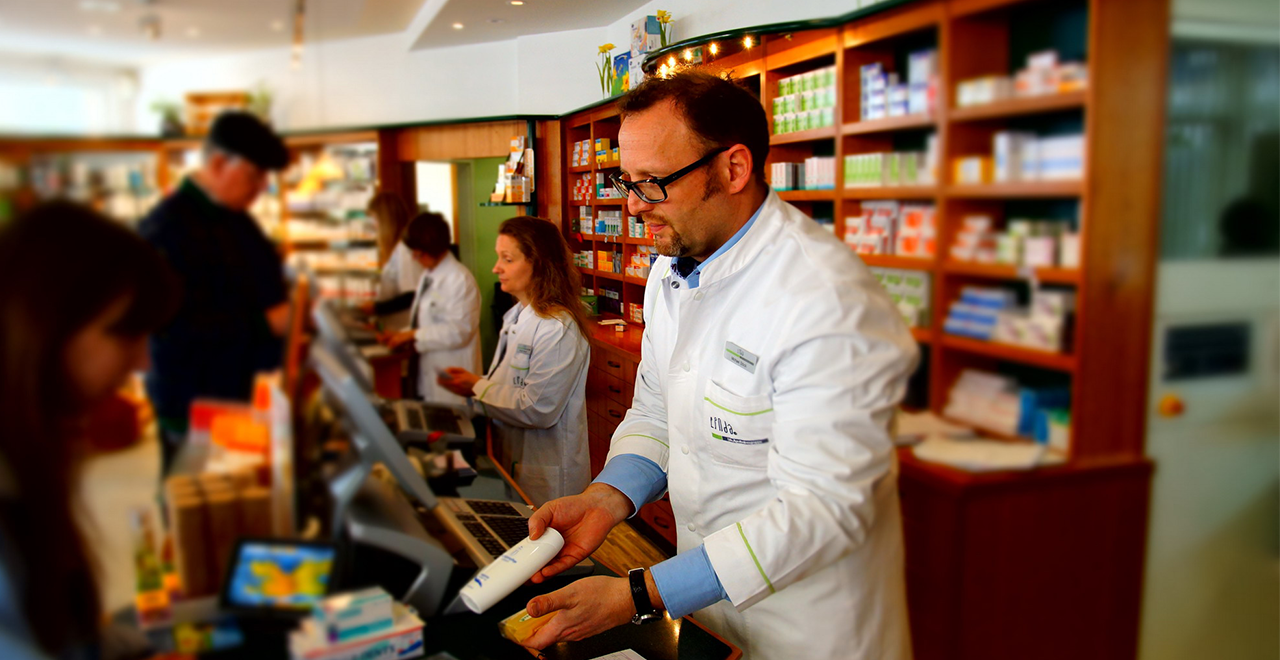 Apotheker im weißen Kittel berät Kundin an der Kasse, weitere Mitarbeiter bedienen andere Kunden im Hintergrund.