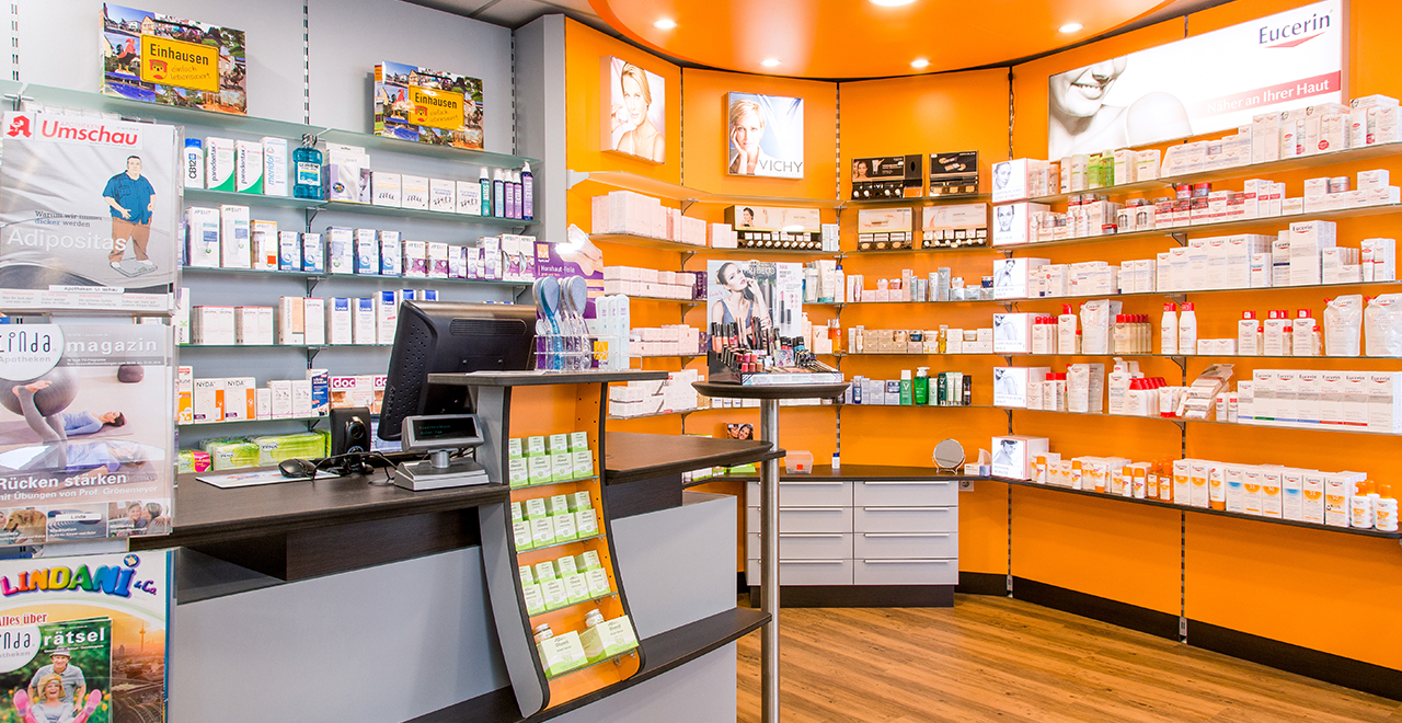 Innenaufnahme einer Apotheke mit einem Empfangstresen, auf dem Medikamente und Pflegeprodukte ausgelegt sind. Im Hintergrund sind weitere Regale sichtbar, die verschiedene Gesundheitsprodukte anbieten.