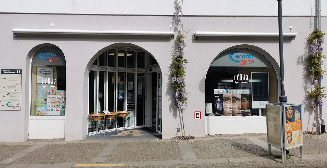 Außenansicht einer Apotheke mit drei Rundbogenfenstern. In der Mitte ist der Eingang mit geöffneten Glastüren. Links und rechts sind Schaufenster mit Werbeplakaten. Über dem Eingang steht ‚Central Apotheke‘ und ‚Linda Apotheken‘. Vor dem Eingang stehen Körbe mit Waren. An der Fassade ranken lila Blütenpflanzen hoch.