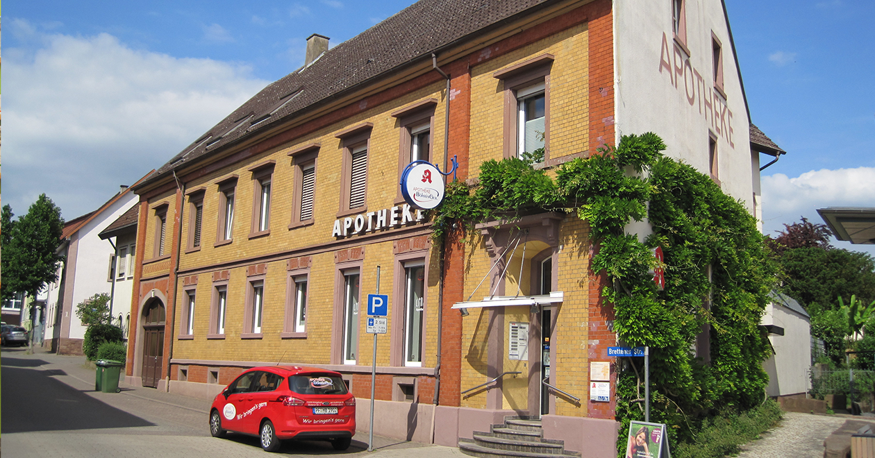 Das Bild zeigt den Verkaufsraum einer Apotheke. Regale mit Medikamenten und Gesundheitsprodukten säumen die Wände. In der Mitte befinden sich Präsentationstische mit weiteren Produkten. Der Boden ist in Holzoptik gehalten, die Atmosphäre ist hell und ordentlich.