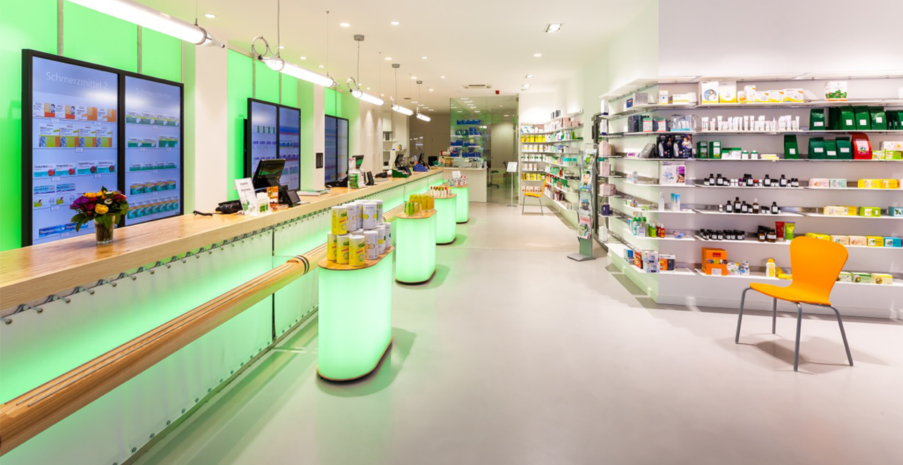 Moderner Verkaufsbereich einer Apotheke mit beleuchtetem, grün akzentuiertem HV-Tisch und mehreren digitalen Bildschirmen zur Produktinformation. Rechts befinden sich übersichtlich angeordnete Regale mit freiverkäuflichen Arzneimitteln, Kosmetik und Gesundheitsprodukten. Im Vordergrund steht ein einzelner orangefarbener Stuhl.