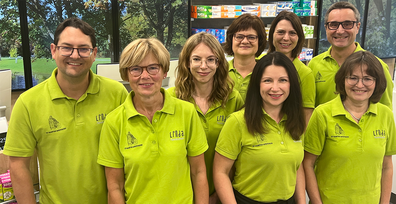 Team der St. Martin Apotheke in grünen Polo-Shirts, lächelnd in der Offizin stehend.