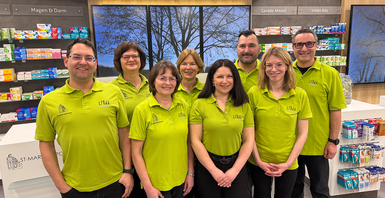 Team der St. Martin Apotheke in grünen Polo-Shirts, lächelnd in der Offizin stehend.