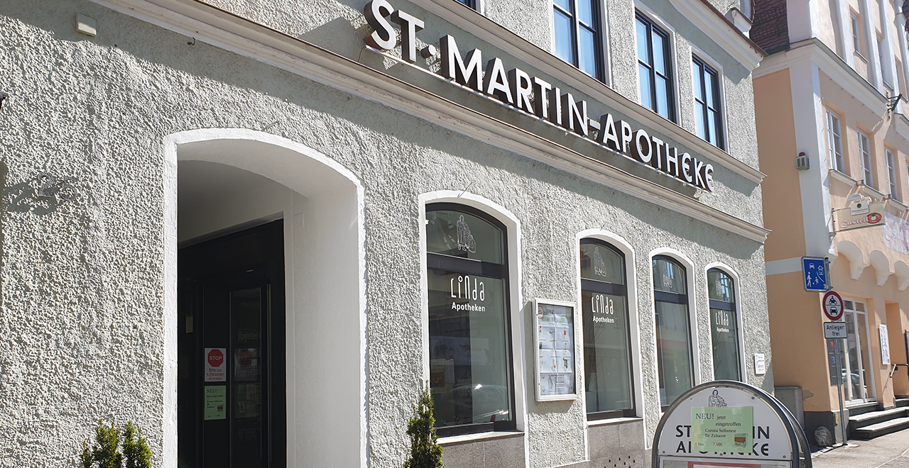 Außenansicht der St. Martin Apotheke mit heller Putzfassade und Rundbogenfenstern. Über der Tür steht „ST. MARTIN-APOTHEKE“. Auf den Fenstern sind das Linda-Logo und Apothekeninformationen sichtbar. Ein Aufsteller informiert über Covid-Impfungen.