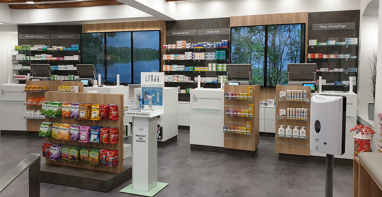 Moderner Innenraum der St. Martin Apotheke mit weißen und holzfarbenen Einrichtungselementen. Vor den großen Bildschirmen mit Naturmotiven befinden sich Kassentresen. Regale mit Medikamenten, Hygieneartikeln und Bonbons sind sichtbar. Ein Desinfektionsspender steht im Vordergrund.