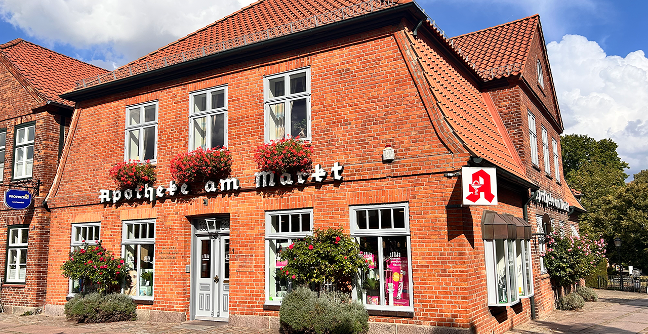 Außenansicht eines historischen roten Backsteingebäudes mit der Aufschrift „Apotheke am Markt“. Das Gebäude hat weiße Fensterrahmen, rote Dachziegel und Blumenkästen mit roten Blumen an den Fenstern. Rechts oben ist das bekannte rote Apotheken-A zu sehen. Im Schaufenster und an der Tür befinden sich LINDA-Logos.
