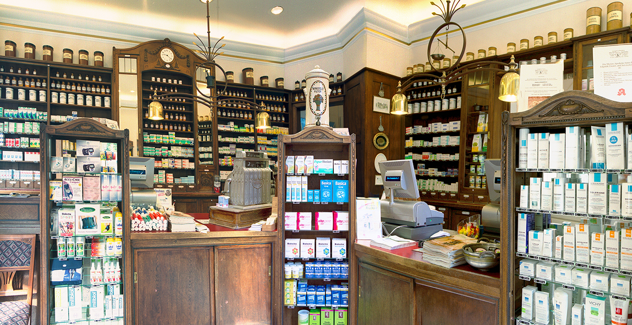 Innenansicht einer traditionellen Apotheke mit dunklen Holzregalen, alten Apothekenschränken, Glasgefäßen und moderner Kassentechnik.