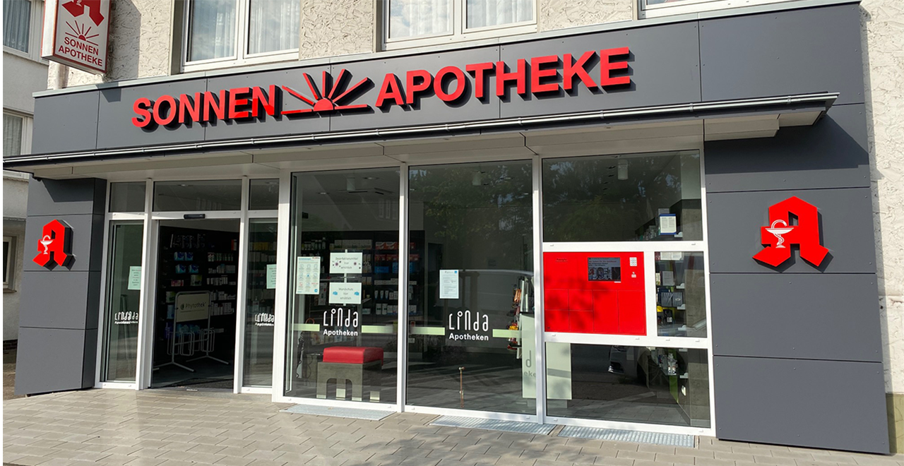 Vorderansicht der Sonnen-Apotheke mit moderner, grauer Fassade. Der Apothekenname „SONNEN APOTHEKE“ ist in großen roten Buchstaben über dem Eingang angebracht, wobei das Wort „Sonnen“ von einer stilisierten roten Sonne begleitet wird. Zwei rote Apotheken-A-Logos sind links und rechts an der Fassade zu sehen. Die Glasfront zeigt das Logo der Linda Apotheken. Rechts neben dem Eingang befindet sich ein roter Abholautomat für Medikamente.