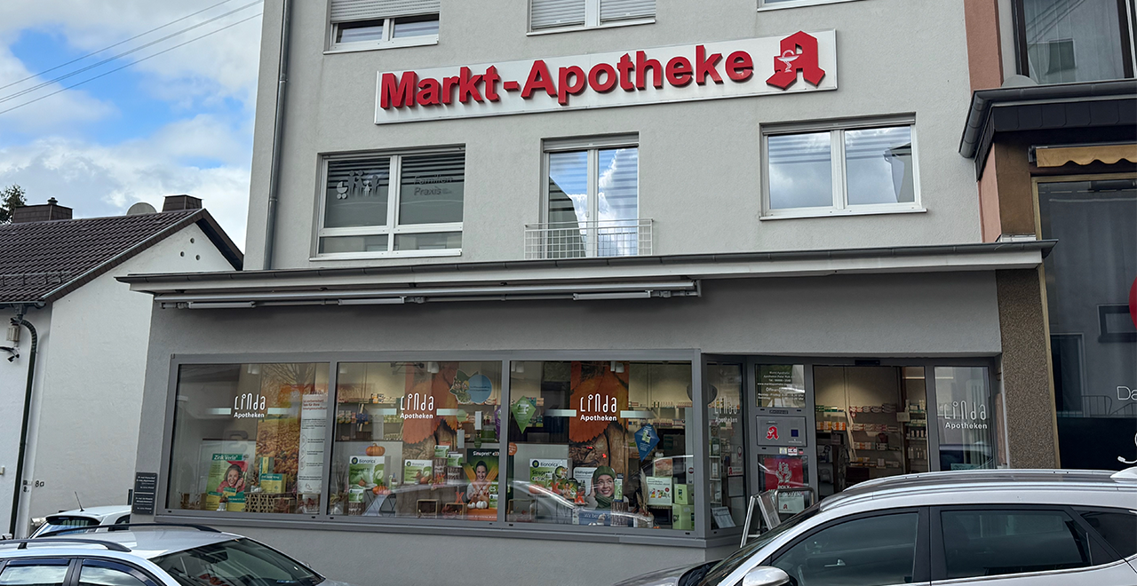 Blick auf den Eingang der Markt-Apotheke, einer Linda Apotheke in einem grauen Gebäude. Die Fenster sind groß und zeigen eine herbstlich dekorierte Auslage mit Kürbissen und Gesundheitsprodukten. Der Eingang ist barrierefrei mit einer Rampe. Über der Tür steht „Linda Apotheken“. Ein Aufsteller mit Angeboten steht draußen.