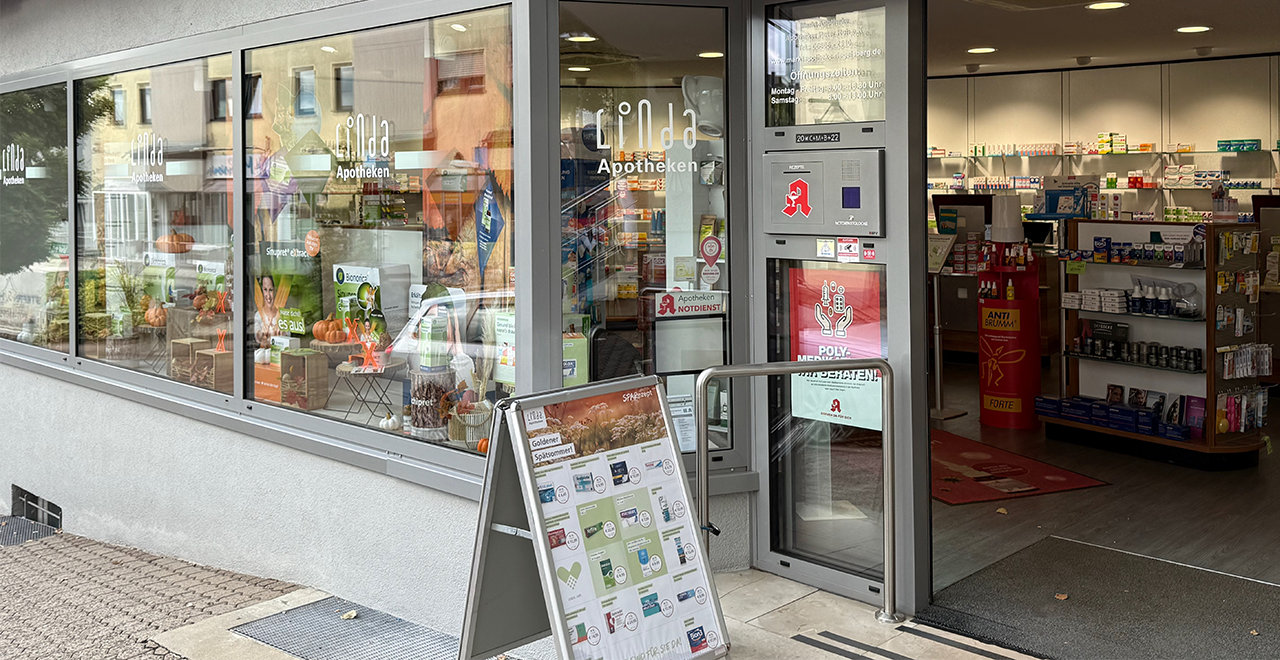 Vorderansicht der Apotheke mit einem weißen Kleinwagen, der „Markt-Apotheke Riegelsberg – wir bringen’s“ in blauer und roter Schrift trägt. Das Auto steht direkt vor dem Schaufenster, in dem zahlreiche Gesundheitsprodukte ausgestellt sind. Rechts der barrierefreie Eingang mit Glasfront.