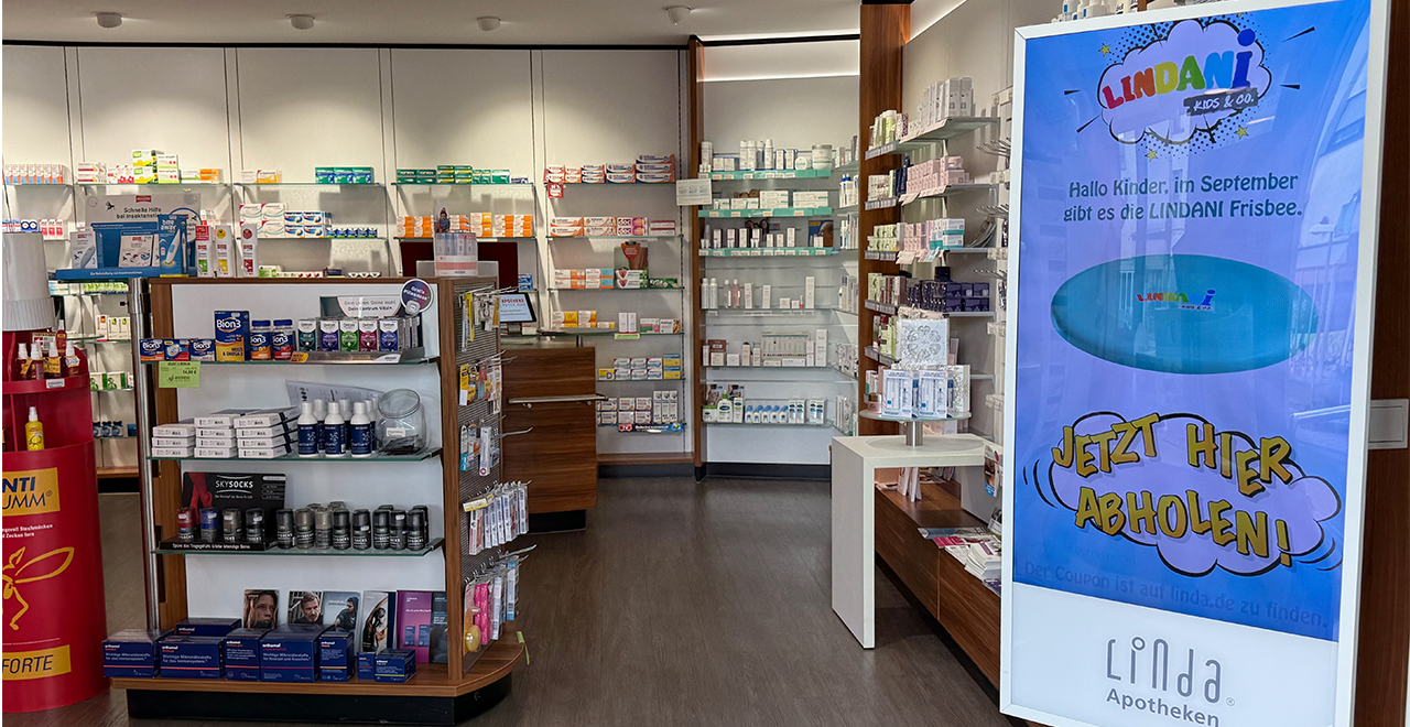 Innenansicht der Apotheke mit zwei modernen Holztresen, Regalen voller Medikamente an der Rückwand und einem runden Verkaufsständer mit Wärmepflastern in der Mitte. Links im Bild ein Desinfektionsspender mit dem Hinweis „Hier bitte Hände desinfizieren“. Die Atmosphäre ist hell und ordentlich.