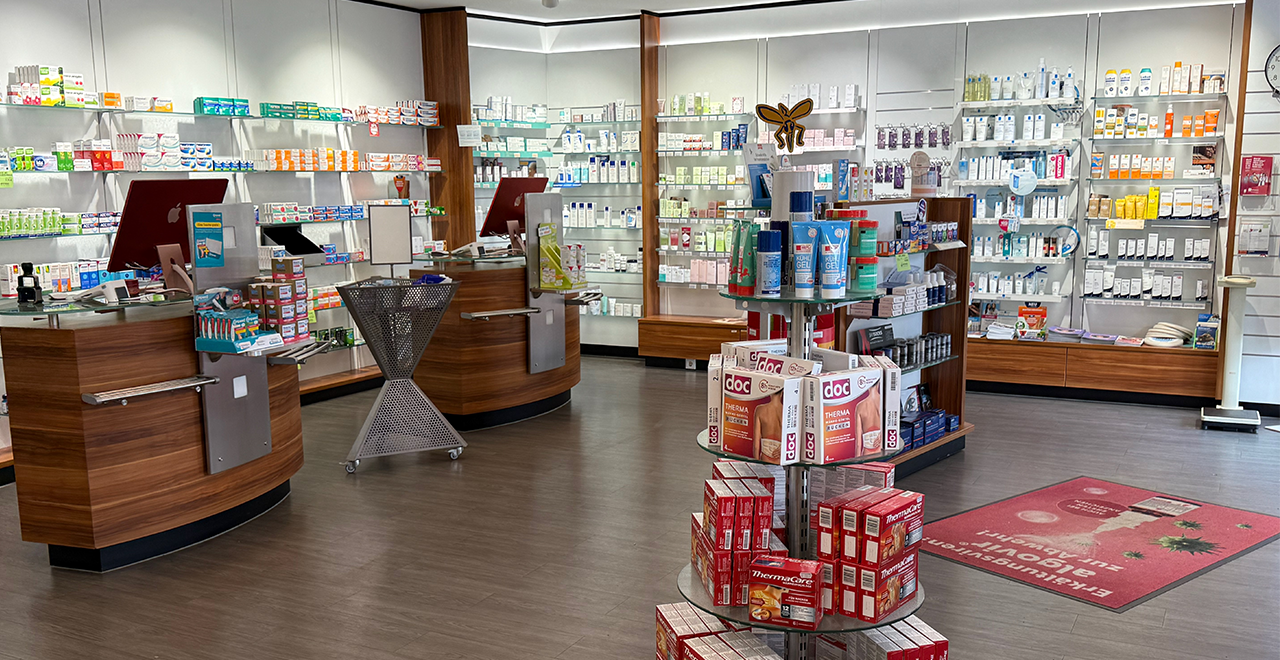 Innenaufnahme der Apotheke mit Fokus auf einen Aufsteller für eine Kinderaktion. Das große Plakat zeigt eine blaue Frisbee mit dem Text „LINDANI Kids & Co. – Jetzt hier abholen!“ im Comic-Stil. Im Hintergrund sind Kosmetik- und Gesundheitsprodukte in Regalen sowie eine Verkaufstheke zu sehen.