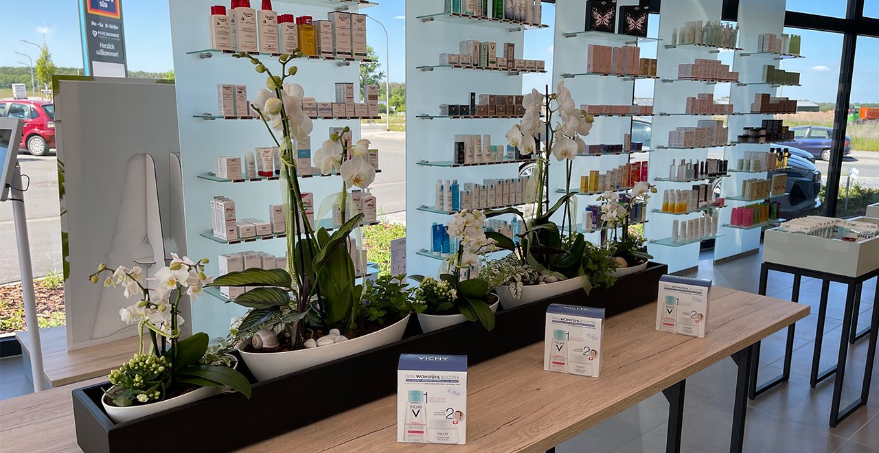 Tisch mit weißen Orchideen und grünen Pflanzen in der Mitte einer hellen Apotheke. Dahinter moderne Glasregale mit Kosmetikprodukten, durch die man nach draußen auf den Parkplatz und die Straße sehen kann.