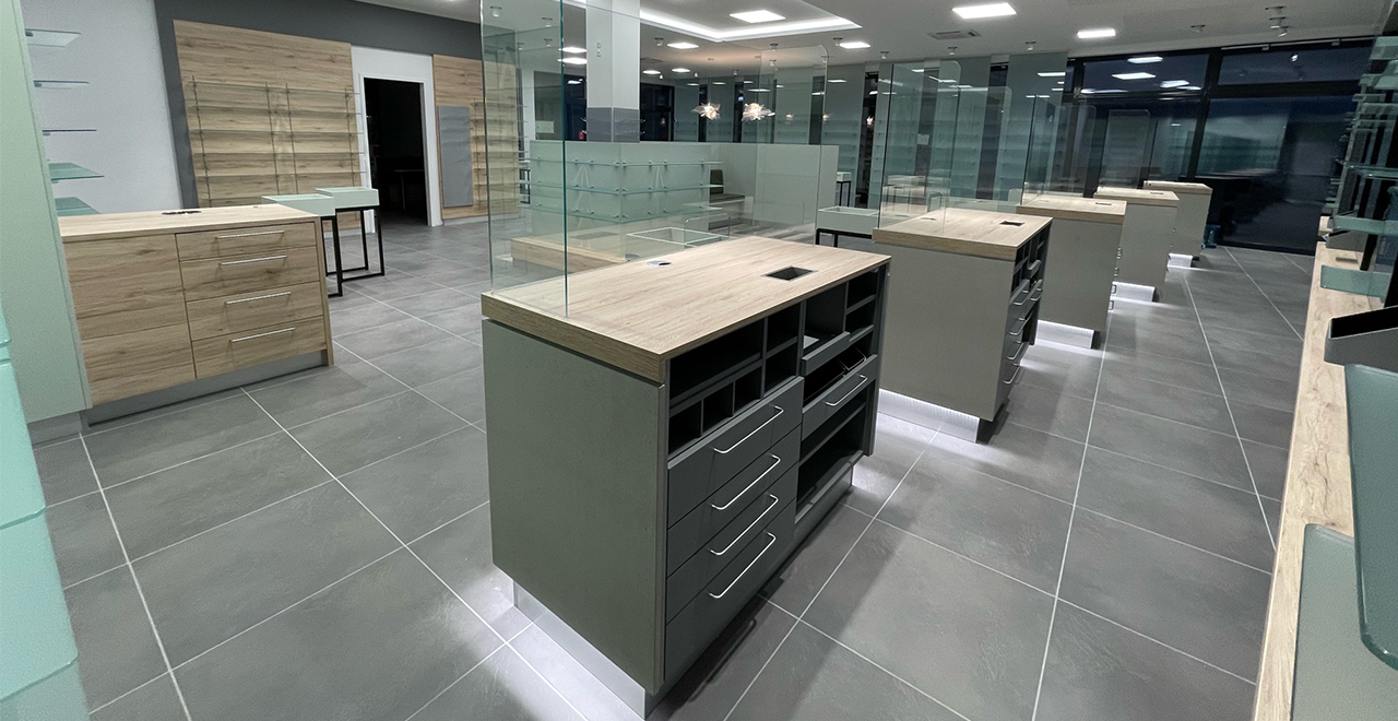 Leerer Innenraum einer modernen Apotheke mit mehreren Bedienungstheken aus Holz und Glas. Die Theken stehen auf einem grauen Fliesenboden, Wände und Regale sind noch nicht mit Produkten bestückt.

