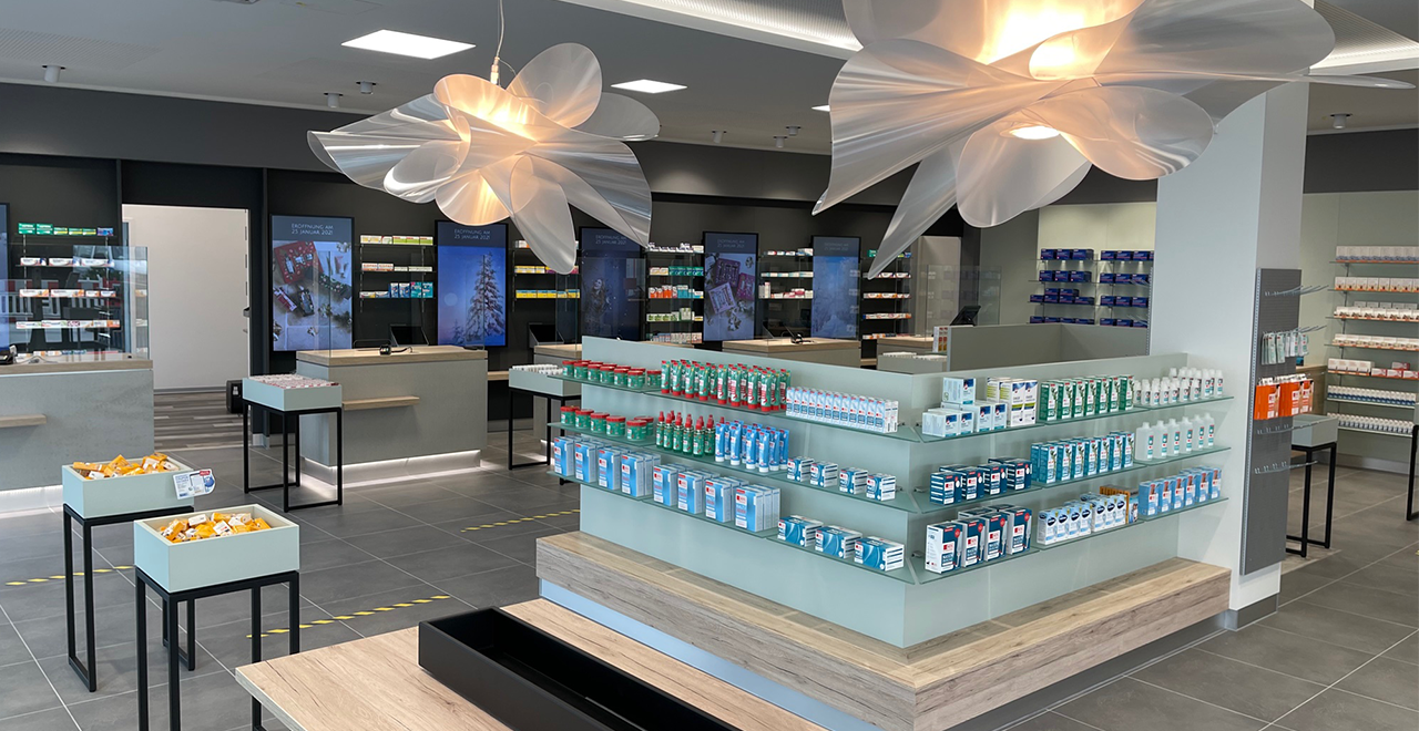 Moderne, helle Apotheke mit eleganter Innenausstattung. In der Mitte stehen Regale mit Medikamenten, an den Wänden befinden sich digitale Bildschirme und weitere Produktregale. Zwei große, kunstvolle Deckenlampen in Blumenform hängen von der Decke.