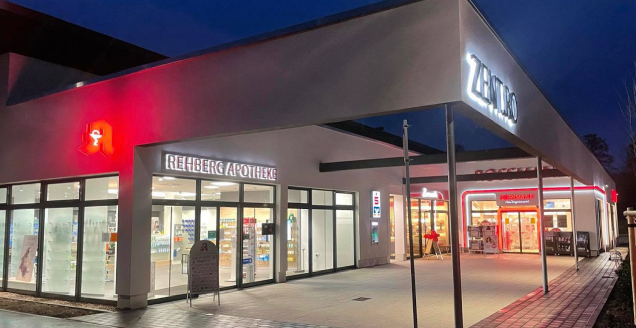 Apothekengebäude bei Nacht mit beleuchtetem Schriftzug „Rehberg Apotheke“ und rotem Apothekenzeichen. Rechts daneben befindet sich eine hell erleuchtete Filiale der Drogeriekette Rossmann. Das Gebäude hat eine moderne Glasfassade und einen überdachten Eingangsbereich.